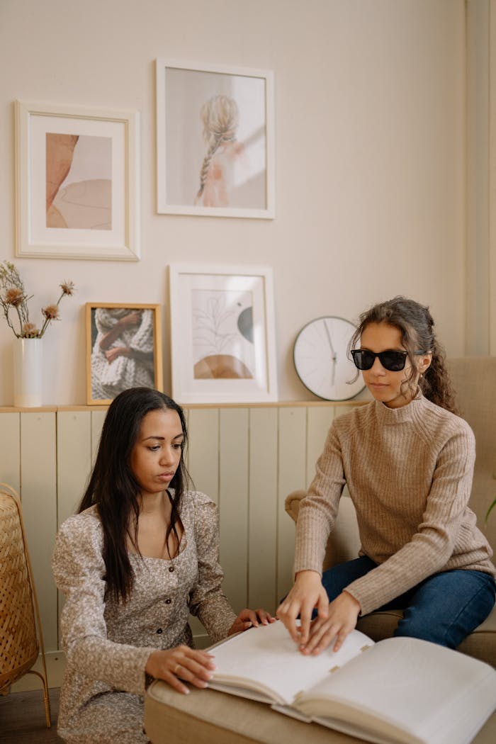 Two women reading a Braille book indoors, one with sunglasses, fostering inclusivity.